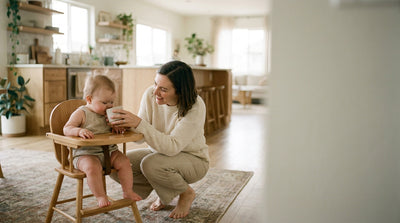 A mother steadying a small open cup for her six month old baby in a high chair to take a first sip of water