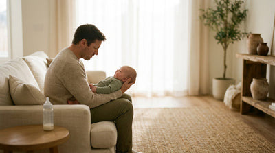 A father sitting on a sofa holding his baby along one forearm, a bottle on the table beside him