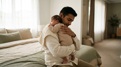 A father burping his baby over his shoulder with a muslin cloth, one hand steady on the baby's back