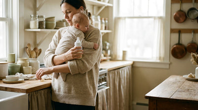 A mother holding her baby on one shoulder while testing the temperature of a bottle taken from the fridge