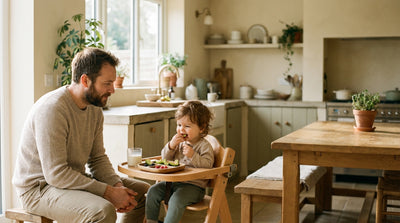 A father sitting beside his one year old, who is self-feeding finger foods in a high chair with a small open cup of milk