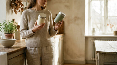 A parent in a kitchen holding two different tins of baby formula, one in each hand, comparing them