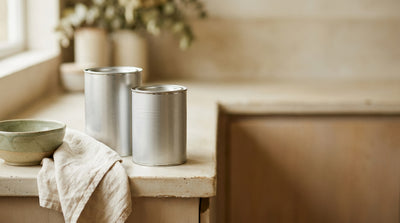 Two tins of baby formula standing side by side on a kitchen counter with a linen cloth and bowl