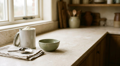 An open tin of baby formula with a measuring scoop, linen cloth and ceramic bowl on a clean kitchen counter