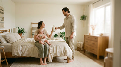 One parent handing a freshly made bottle to the other, who is holding their baby on the edge of the bed