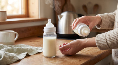 A parent testing a few drops of formula on the inside of their wrist beside a filled baby bottle on a kitchen counter.