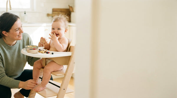 8 month old baby self-feeding in a high chair while a parent sits nearby